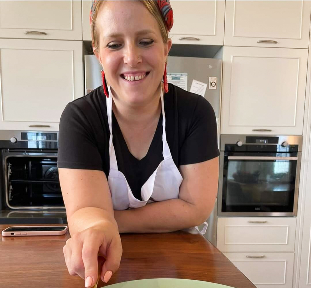 A woman leaning on a table staring at a plate of food she cooked. She is wearing an apron and smiling.