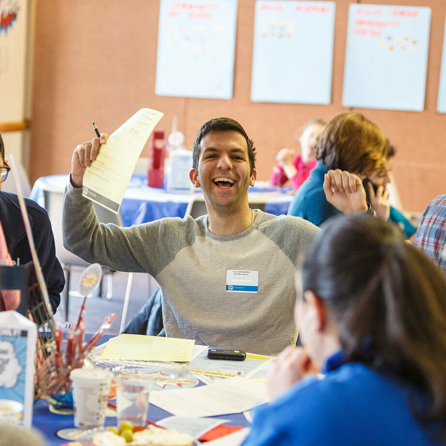 A young man holding his arms up. There is a piece of paper in his right hand and he is smiling.