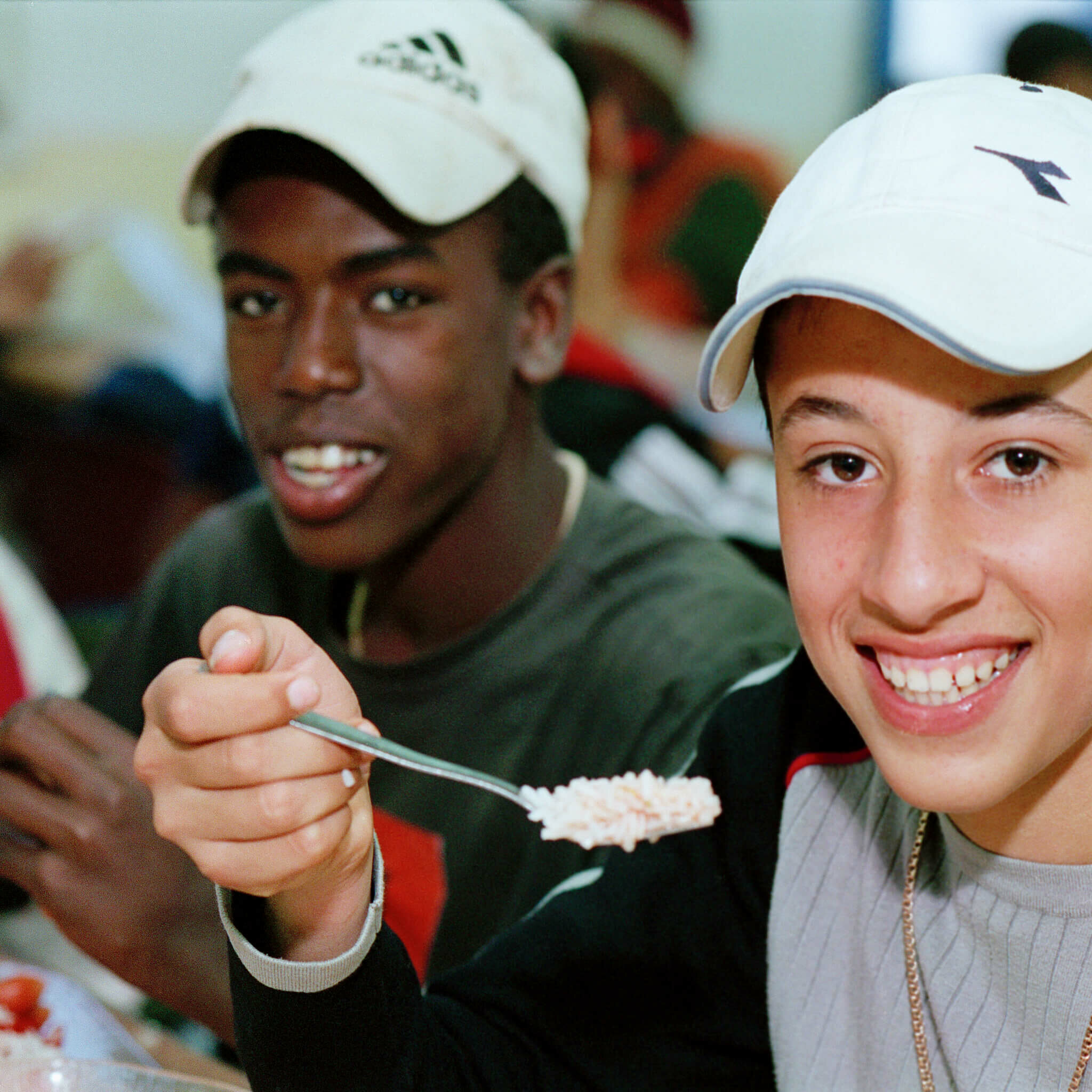 Two teens sitting at a table smiling. The one has a fork with food on it in midair.